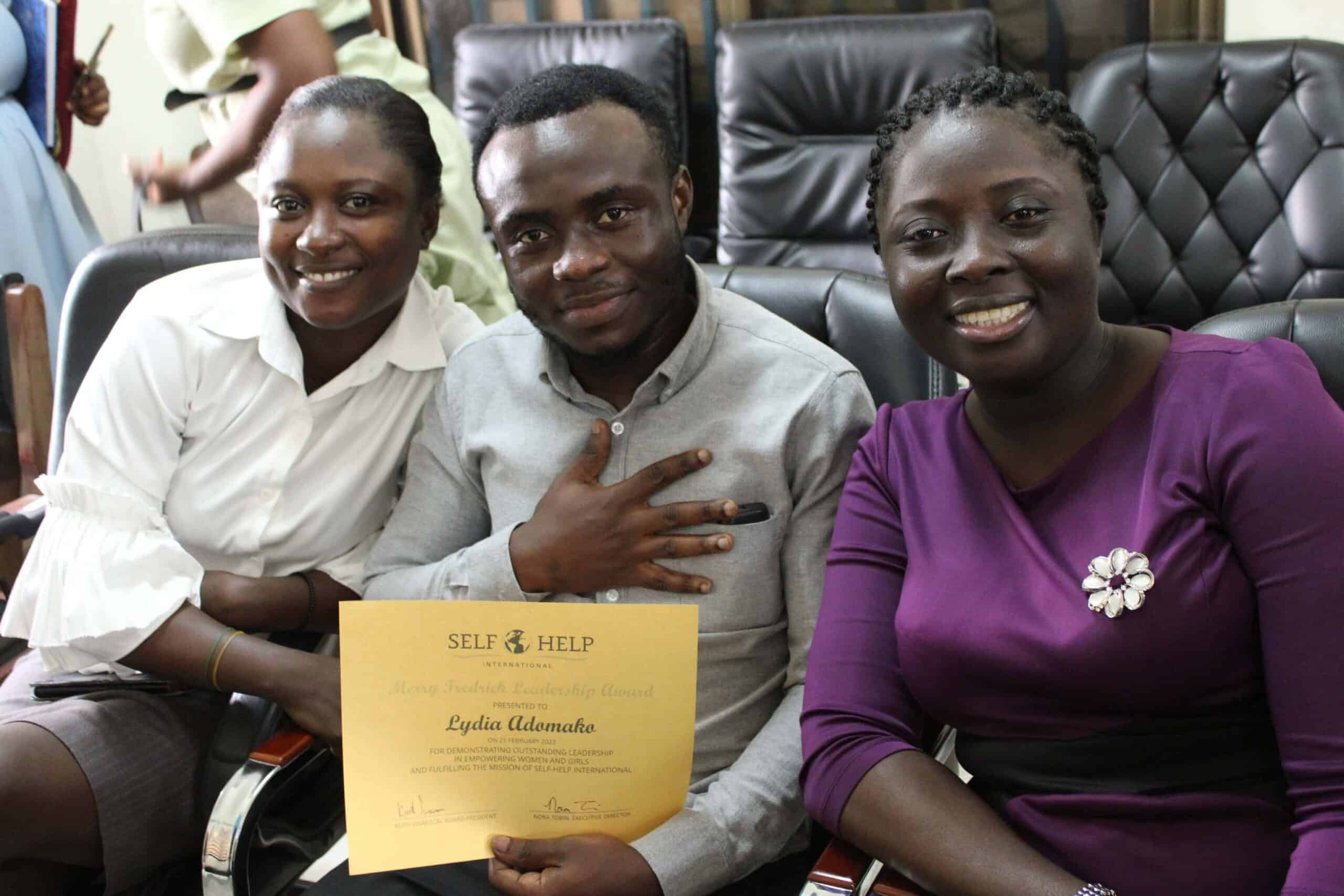 Three Ghanaian staff members are seated and posing for a photo. Lydia, wearing a purple dress, smiles on the right. To her left are colleagues Chris and Florence, also smiling. Chris is holding the yellow certificate recognizing Lydia's leadership.