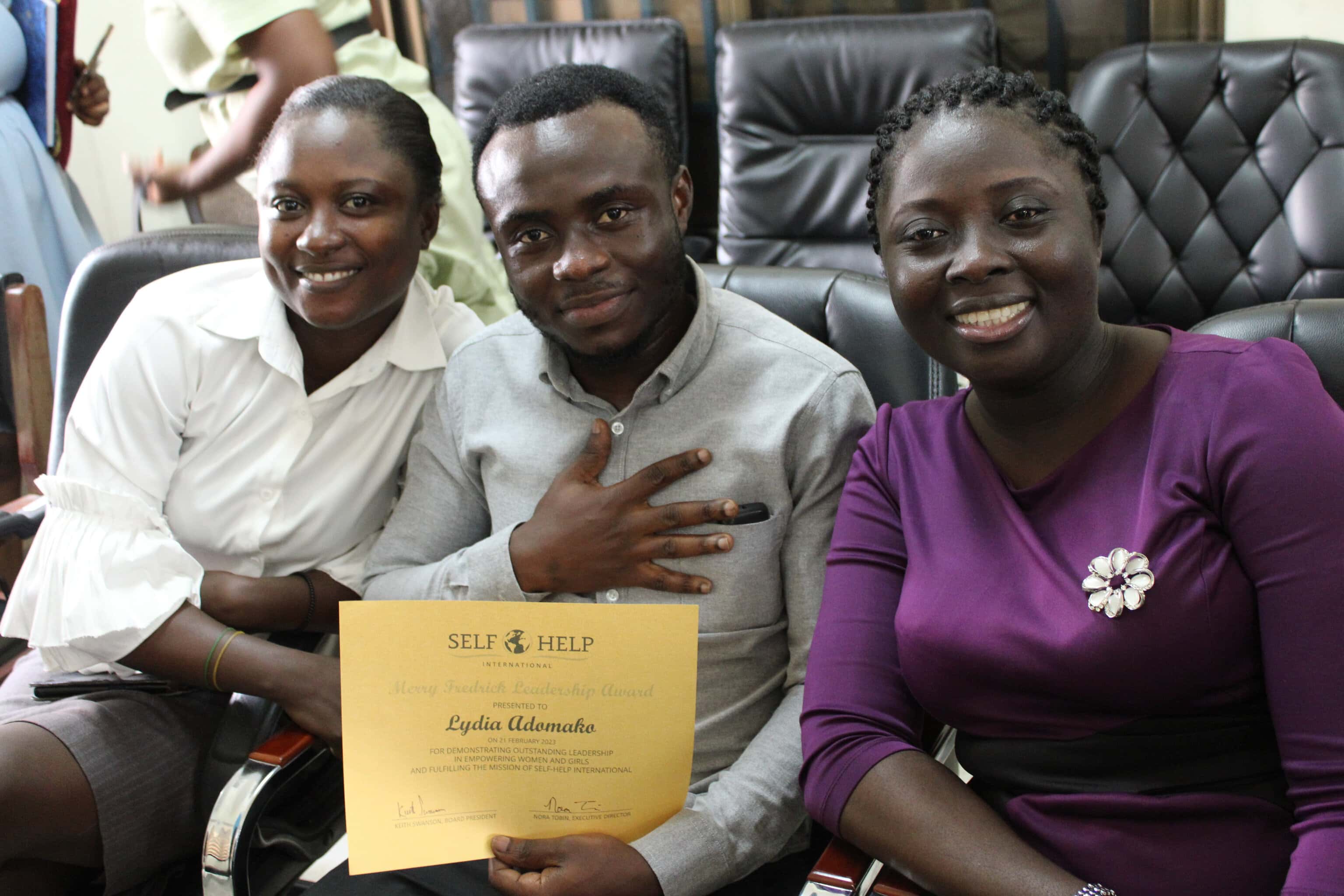 Three Ghanaian staff members are seated and posing for a photo. Lydia, wearing a purple dress, smiles on the right. To her left are colleagues Chris and Florence, also smiling. Chris is holding the yellow certificate recognizing Lydia's leadership.